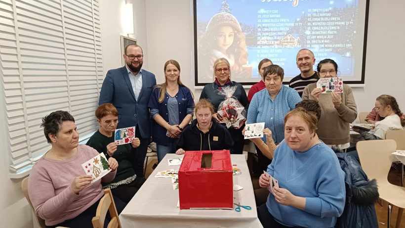 Mieszkańcy i personel Domu Pomocy Społecznej w Lubzinie pozują do grupowego zdjęcia, dumnie prezentując ręcznie robione kartki świąteczne wykonane podczas warsztatów.