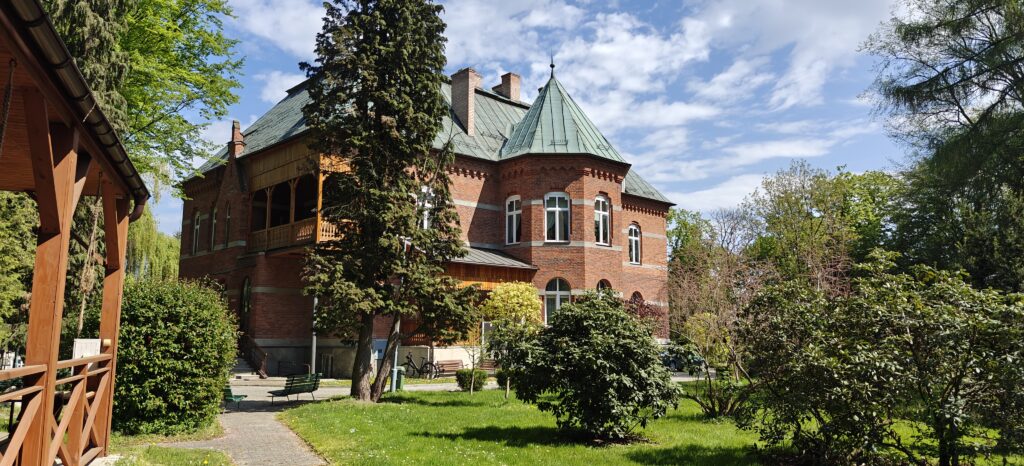 Widok na budynek Domu Pomocy Społecznej w Lubzinie od strony parku. Historyczny gmach z czerwonej cegły otoczony jest zadbanym trawnikiem, drzewami, alejką spacerową i drewnianą altaną.
