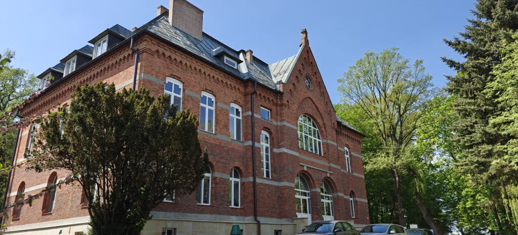 Główny budynek administracji Domu Pomocy Społecznej w Lubzinie widziany z zewnątrz w słoneczny dzień. Historyczny, piętrowy gmach z czerwonej cegły otoczony jest wysokimi, zielonymi drzewami.