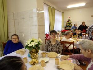 Mieszkańcy Domu Pomocy Społecznej w Lubzinie siedzą przy wspólnym stole podczas świątecznego posiłku. Na stole stoi biała gwiazda betlejemska, w tle widoczna jest choinka.